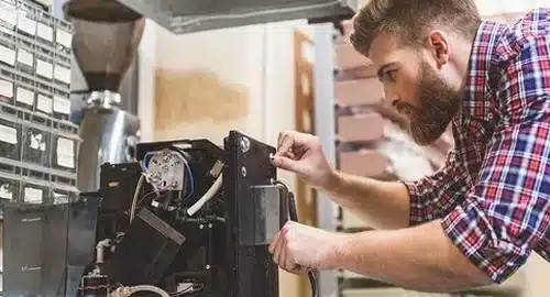 A man with a beard, wearing a plaid shirt, is using a screwdriver to repair or assemble an electronic device with visible wires and components on a workbench in a workshop.
