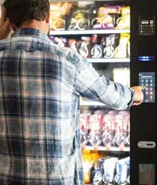 A person in a blue plaid shirt stands in front of a vending machine, using the keypad to make a selection. The machine is filled with various snacks and drinks.