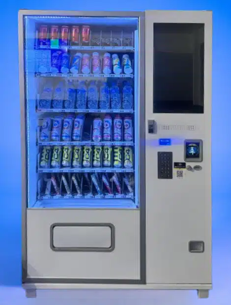 A brightly lit vending machine displays assorted energy drinks and sodas behind a glass front. The machine is white with a digital payment terminal and keypad on the right side, set against a blue background.