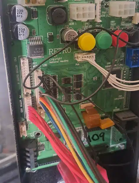 Close-up of a green circuit board labeled "RETRO" with various colored wires connected, large capacitors, buttons (yellow, green, red), and electronic components inside a vending machine.