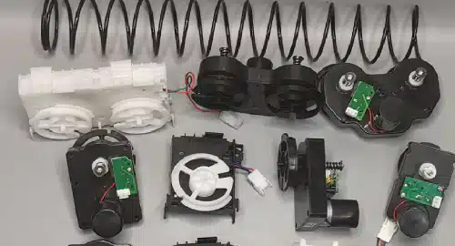 Assorted mechanical and electronic components, including gears, circuit boards, and coiled springs, are arranged on a flat surface. The parts are primarily black and white with visible wiring.